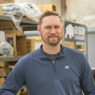 A man in a warehouse wearing a blue quarter zip shirt looking at the camera and smiling