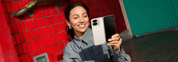 Bir kişi, elindeki porselen renk Pixel 9 Pro Fold telefona bakıp gülümsüyor. Arka planda kırmızı fayans kaplı bir duvar var.