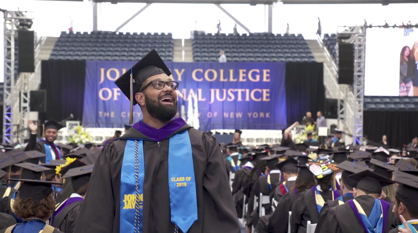 Excited John Jay College student dressed up in cap and gown, standing in auditorium at graduation ceremony