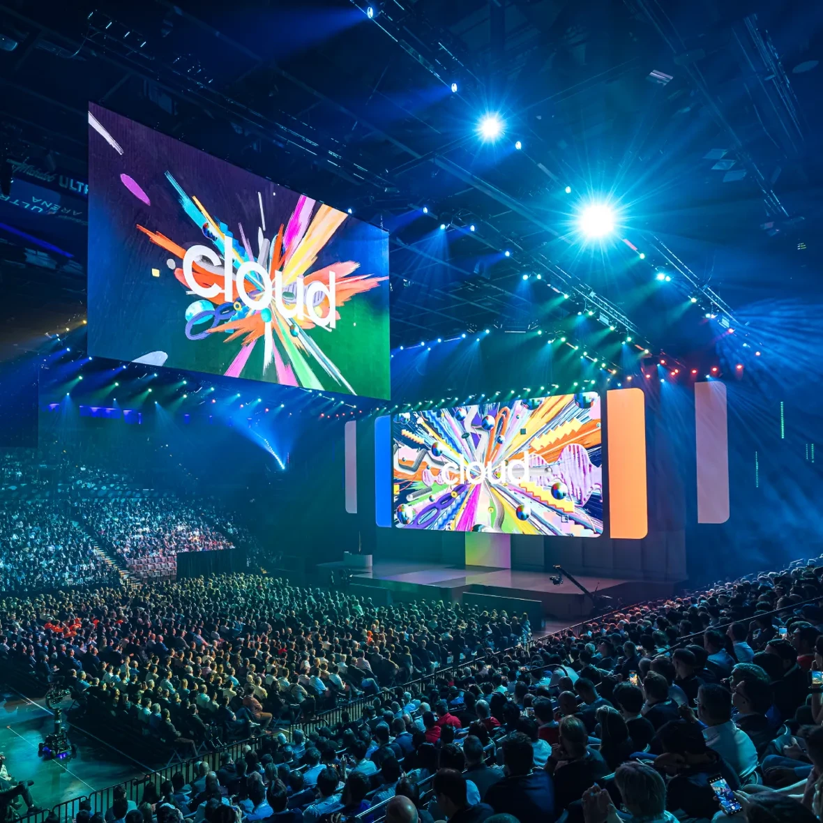 Large indoor event with a packed audience facing a stage with two large screens displaying colorful abstract graphics