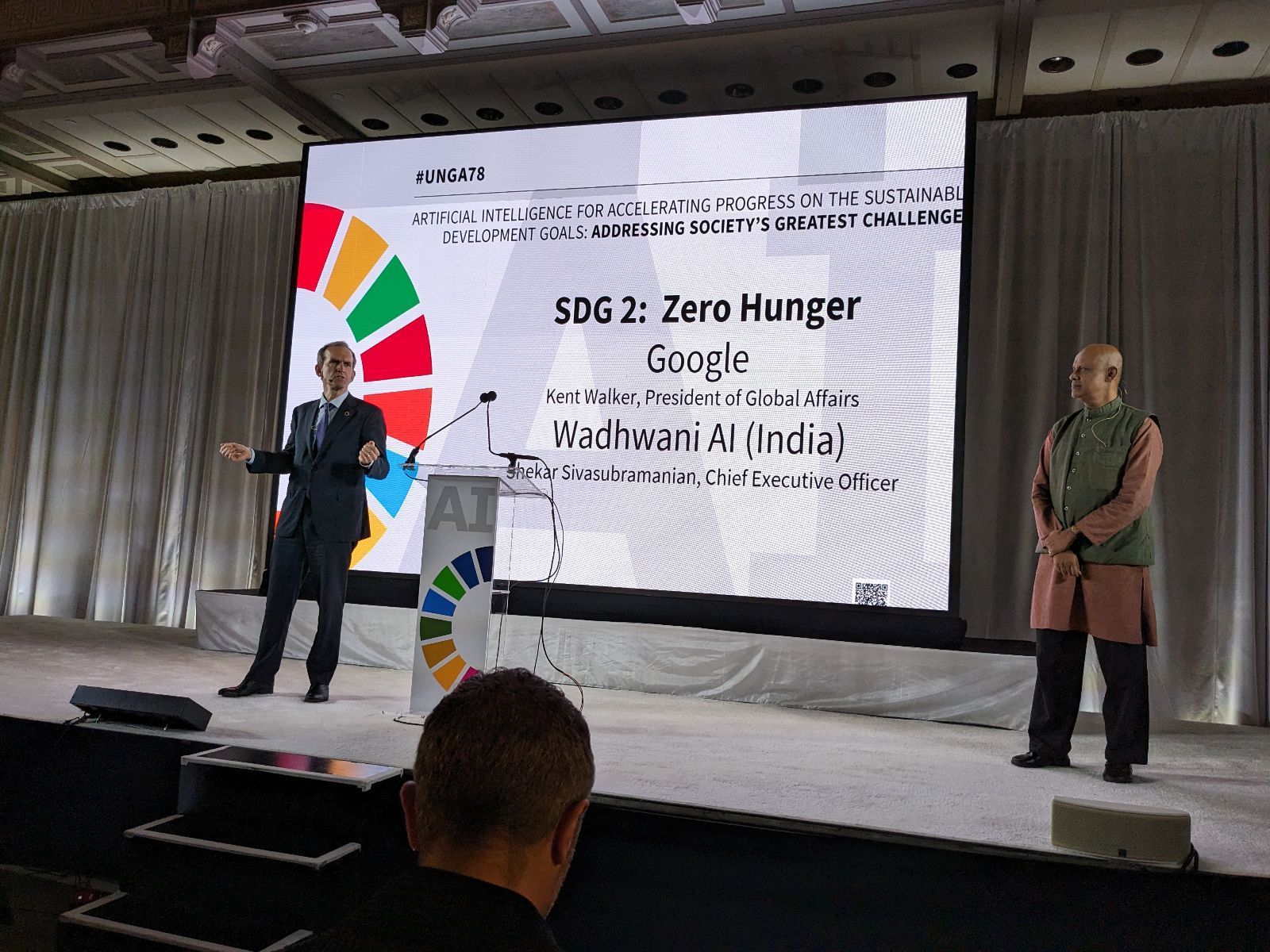 Man in dark suit speaking on stage, standing in front of a projection screen with a multicolor wheel and text on screen.