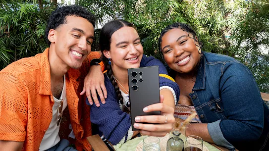Three people smile for a selfie as the middle person captures the moment with an Android-compatible phone.