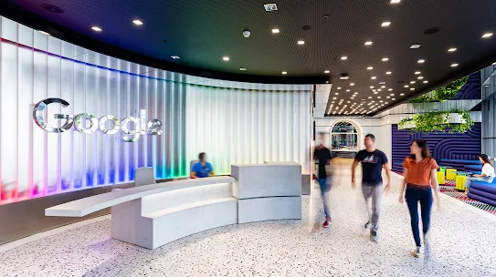Red, blue and green lighting illuminate a curved wall with a silver Google logo. In front, a person sits behind a white desk, with three people passing by.