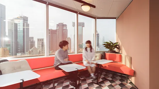 Two people sit on a red couch in front of windows.